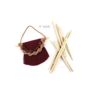 May include: A 6 inch macrame wall hanging with a burgundy and brown color scheme. The hanging features a half-circle design with wooden beads and a fringe. The hanging is attached to a wooden dowel.