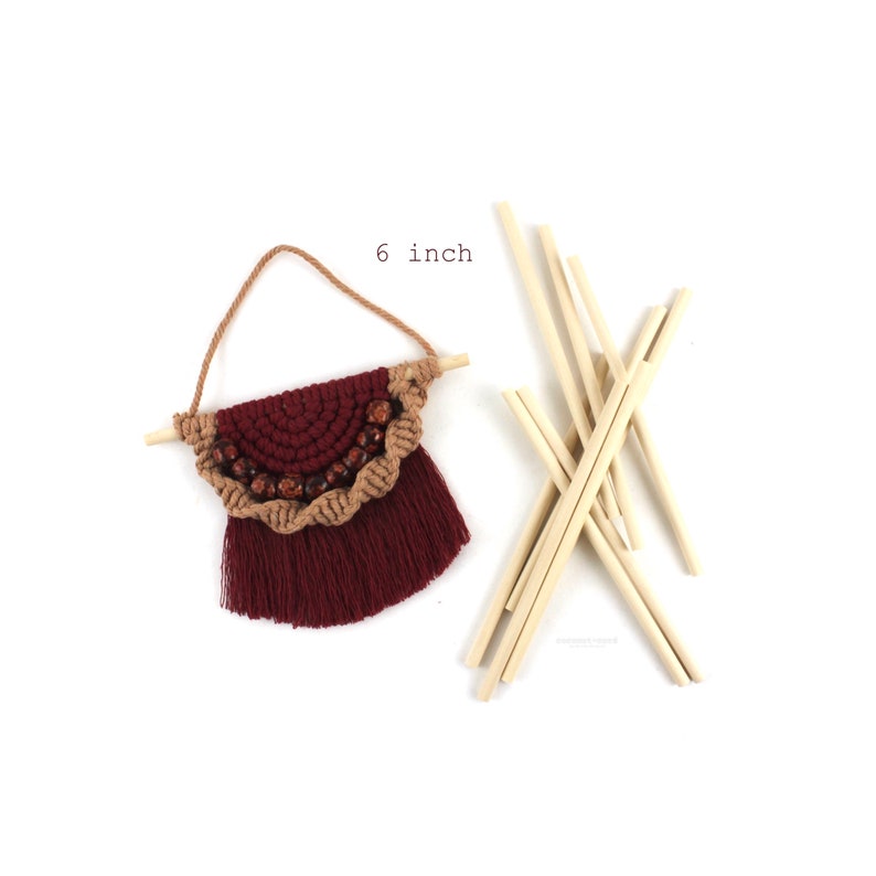 May include: A 6 inch macrame wall hanging with a burgundy and brown color scheme. The hanging features a half-circle design with wooden beads and a fringe. The hanging is attached to a wooden dowel.