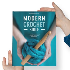 Peut inclure: Un livre bleu turquoise intitulé "Modern Crochet Bible" de Sarah Shrimpton. La couverture présente une paire de mains tenant une pelote de laine bleu turquoise et un crochet en bois.