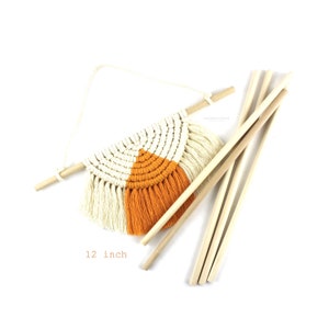 May include: A macrame wall hanging with a white and orange design. The hanging is 12 inches wide and comes with wooden dowels for hanging.