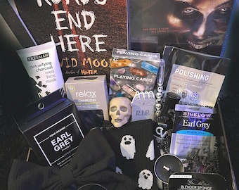 May include: A Halloween-themed gift basket with a black and white book titled "All Roads End Here", a Blu-ray disc of the movie "The Purge", a black face mask, a skull-shaped soap dispenser, a box of Earl Grey tea, a black bow, a vanilla scented candle, a black and white ghost-patterned sock, and a black and white playing card deck.