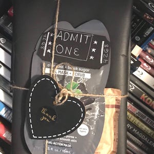 May include: A black gift box wrapped with twine and a black heart-shaped tag that says "thank you". The box is topped with a black and gray face mask with the text "charcoal + black sugar L MASK + CRUB" and a white circle that says "all skin types". The box is also decorated with a black and white ticket that says "ADMIT ONE" and "892th".