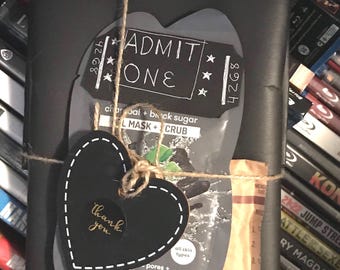 Puede incluir: Una caja de regalo negra envuelta con cuerda y una etiqueta negra en forma de corazón que dice "gracias". La caja está coronada con una mascarilla facial negra y gris con el texto "charcoal + black sugar L MASK + CRUB" y un círculo blanco que dice "todos los tipos de piel". La caja también está decorada con un boleto negro y blanco que dice "ADMIT ONE" y "892th".