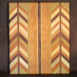 Op de afbeelding: Een vierkante houten snijplank met een decoratief visgraatpatroon. De plank heeft een verscheidenheid aan houttinten, waaronder licht- en donkerbruin, oranje en groen. Het centrale gedeelte heeft verticale strepen.