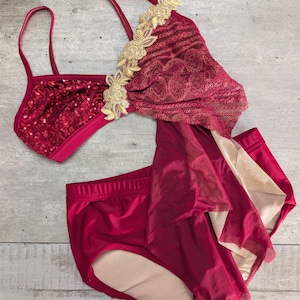 May include: A two-piece dance costume featuring a deep red sequined crop top with thin straps and gold floral appliqué detailing. The matching high-waisted bottoms are deep red with a layered sheer fabric skirt overlay and a beige lining.
