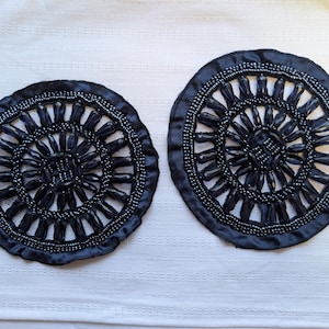 May include: Two dark blue, circular decorative pieces with intricate beaded designs. Each piece features a central beaded square, surrounded by radiating beaded elements and a beaded border. The pieces are placed on a white surface.