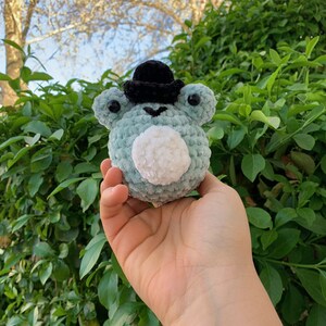 May include: A small, crocheted frog wearing a black top hat. The frog is light green with a white fluffy belly.