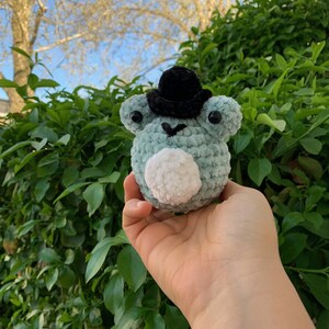 May include: A small, crocheted frog wearing a black hat. The frog is light blue and white with black eyes. The frog is being held in a hand.