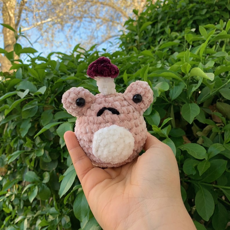 May include: A pink crocheted frog with a purple mushroom on its head. The frog has black eyes and a white belly.