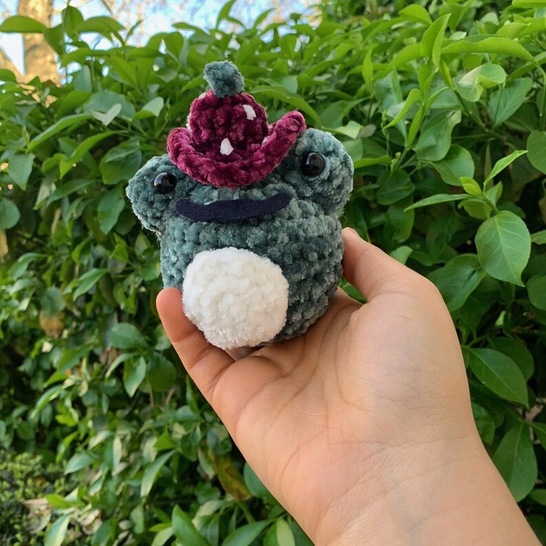 May include: A green crocheted frog wearing a burgundy velvet mushroom hat with white polka dots. The frog has white fuzzy eyes and a white fuzzy belly.