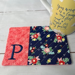 May include: A pink and blue floral fabric coaster with a navy blue letter P embroidered on the pink side. The coaster is quilted and has a white background with small white dots. The coaster is sitting on a white surface. The text on the mug reads "Your word is a lamp to my feet and a light for my path Psalm 119:105"