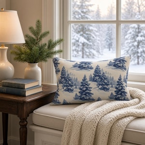 May include: A rectangular throw pillow featuring a blue and white toile-style pattern of evergreen trees in a snowy landscape. The pillow is positioned on a window seat next to a cream-colored lamp, a small artificial pine tree, and books.