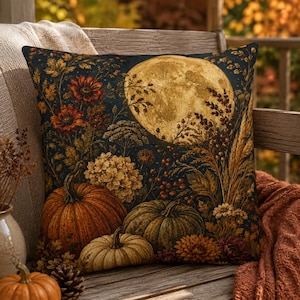 Könnte beinhalten: Ein quadratisches Dekokissen mit dunkelblauem Hintergrund und einem großen, leuchtend gelben Vollmond. Das Motiv zeigt orangefarbene und grüne Kürbisse, herbstliche Blumensträuße, getrocknete Weizenhalme und Blätter in Gold- und Brauntönen.