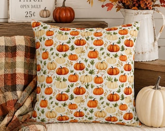 May include: A square throw pillow featuring a repeating pattern of orange and cream-colored pumpkins interspersed with green leaves on a white background. The pillow is displayed alongside a plaid blanket and a white pitcher filled with autumn foliage.
