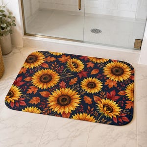 Op de afbeelding: Een rechthoekige badmat met een donkerblauwe achtergrond en een herhalend patroon van levendige gele en oranje zonnebloemen, afgewisseld met dieprode en bruine herfstbladeren en kleine trosjes donkerrode bessen over het oppervlak.