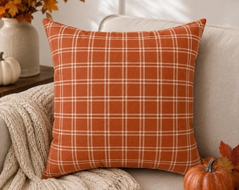 May include: A square throw pillow featuring a burnt orange fabric base with a white grid pattern. The pillow has a clean, structured shape with visible seams along the edges, showcasing a consistent plaid design across the entire front surface.