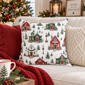 May include: A square throw pillow with a white background featuring a repeating pattern of red and green houses, snow-covered roofs, evergreen trees, and black street lamps. The fabric has a textured weave, and the design depicts a winter village scene.