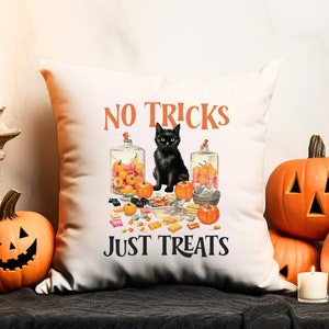 Puede incluir: Coj&iacute;n cuadrado blanco con tem&aacute;tica de Halloween. El coj&iacute;n presenta un gato negro, calabazas y caramelos. El texto en el coj&iacute;n dice "NO TRICKS JUST TREATS" en naranja y negro. Calabazas decorativas y una vela encendida en el fondo.