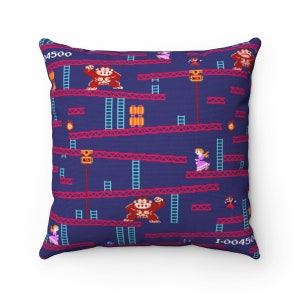 May include: A blue throw pillow with a pixelated pattern of a video game level. The pattern includes a monkey, a princess, and a ladder. The text "1-004500" is printed in the bottom right corner.