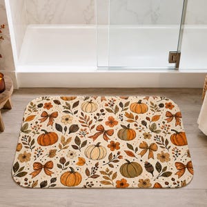 May include: A rectangular bath mat featuring a repeating pattern of orange and cream pumpkins, brown autumn leaves, small flowers, and tied ribbons on a light cream background with rounded corners and a soft, plush texture.