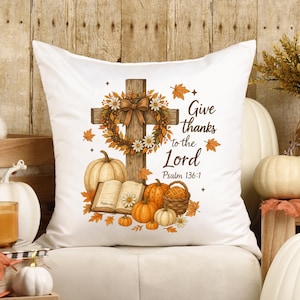 May include: White square cushion with a fall design. The design includes a wooden cross with a floral wreath, pumpkins, and the text "Give thanks to the Lord" from Psalm 136:1. The cushion has a soft, inviting appearance.
