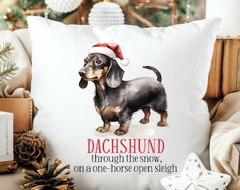 Può includere: Un cuscino bianco con un bassotto nero e marrone che indossa un cappello da Babbo Natale rosso. Il testo sul cuscino recita "Dachshund through the snow, on a one-horse open sleigh."