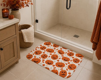 Puede incluir: Una alfombrilla de baño rectangular con un patrón repetitivo de calabazas de Halloween naranjas con diversas expresiones faciales talladas, intercaladas con pequeñas hojas de otoño marrones sobre un fondo blanco con esquinas redondeadas.
