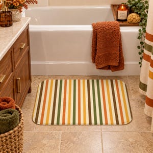 Peut inclure: Un tapis de bain rectangulaire orné d'un motif à rayures verticales dans des tons de brun foncé, vert olive, jaune moutarde, orange et crème. Le tapis présente des coins arrondis et une surface lisse, posé sur un sol de salle de bain carrelé.