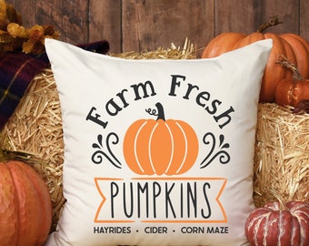 Puede incluir: Un cojín cuadrado blanco con un gráfico central de una calabaza naranja y texto negro que dice Farm Fresh PUMPKINS, con texto más pequeño debajo que indica HAYRIDES, CIDER y CORN MAZE, acentuado con adornos decorativos negros.