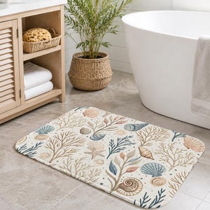 May include: Rectangular bath mat with a cream background and a watercolor design of seashells, coral, and seaweed in shades of blue, tan, and brown. The mat is placed on a tiled floor in a bathroom setting.