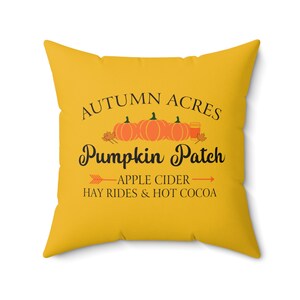 May include: Yellow throw pillow with the text "Autumn Acres Pumpkin Patch" in black. The text is surrounded by a row of orange pumpkins and a cup of hot cocoa. Below the text is a black arrow pointing to the left with the text "Apple Cider" and a black arrow pointing to the right with the text "Hay Rides & Hot Cocoa".
