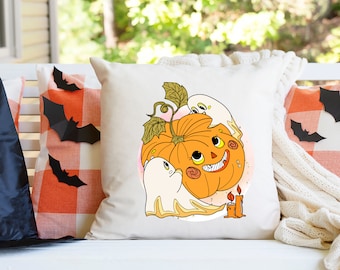 May include: A square off-white throw pillow featuring a central graphic of a smiling orange jack-o'-lantern with green leaves, two white ghosts, and two lit orange candles. The design is rendered in a cartoon style with bold black outlines.