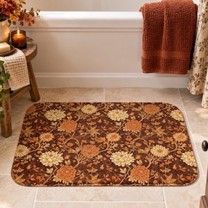 May include: A rectangular bath mat featuring a dark brown background with a repeating pattern of orange and cream-colored chrysanthemum flowers, winding stems, and leaves. The mat has rounded corners and a soft, textured surface finish.