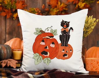 May include: A white square throw pillow featuring a graphic illustration of two orange jack-o'-lanterns with carved faces and green leaves, topped by a black cat wearing an orange and white polka-dot bow tie, centered on the fabric surface.