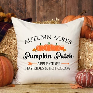 May include: White throw pillow with the text "Autumn Acres Pumpkin Patch" in black cursive font.  The text is surrounded by three orange pumpkins and a cup of hot cocoa.  Below the pumpkins is the text "Apple Cider Hay Rides & Hot Cocoa" in black.