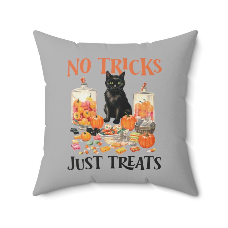 Puede incluir: Un coj&iacute;n cuadrado gris con el texto "NO TRICKS JUST TREATS" en naranja y negro. El coj&iacute;n presenta un gato negro, calabazas y tarros de caramelos, creando un tema de Halloween.