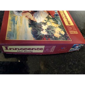 May include: A 750-piece jigsaw puzzle box with a painting of two children in a field of yellow flowers. The box is red with the title "Innocence Get Ready" printed on the front.