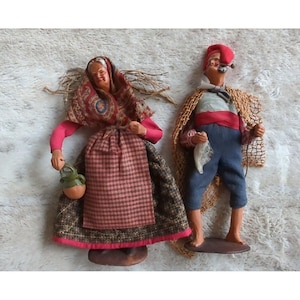 May include: Two vintage figurines of a man and woman dressed in traditional clothing. The woman is wearing a red, black, and white dress with a patterned shawl. The man is wearing a blue and white striped shirt, red pants, and a red hat. Both figurines are holding objects in their hands.