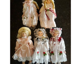 May include: Five porcelain dolls dressed in vintage-inspired dresses with lace trim and floral accents. The dolls have blonde hair and are wearing hats.