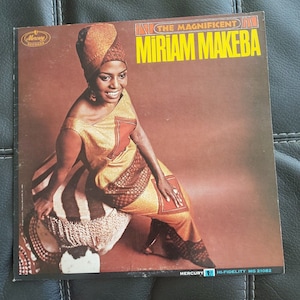 Peut inclure: Une pochette d'album de disque vinyle vintage mettant en vedette Miriam Makeba, une chanteuse, compositrice et militante sud-africaine. La pochette présente une photo de Makeba dans une robe colorée, assise sur un tabouret. Le titre de l'album, "The Magnificent Miriam Makeba", est imprimé en grandes lettres audacieuses en haut de la pochette. L'album est une sortie de Mercury Records.