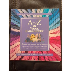 May include: A book titled "A-Z of Wool Embroidery" with a purple cover and a colorful yarn border. The book cover features a small illustration of a sheep surrounded by flowers. The text on the cover reads "Discover the delights of wool embroidery" and "The fourth book in the successful A-Z series."