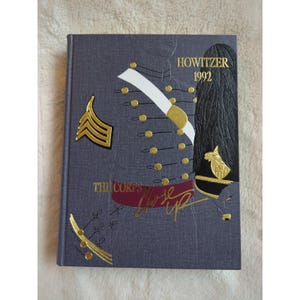 Pode incluir: Um livro encadernado em capa dura cinza com letras douradas. O título é "Howitzer 1992" e o subtítulo é "The Corps' Close Up". A capa apresenta uma ilustração estilizada de um uniforme militar com detalhes dourados.