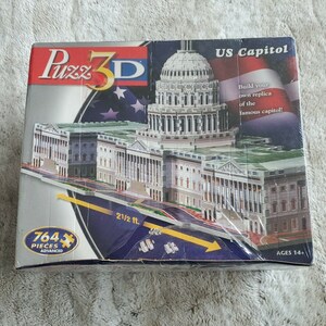 Peut inclure: Un puzzle 3D de 764 pièces représentant le Capitole des États-Unis. La boîte montre une image détaillée du bâtiment avec le texte "US Capitol" et "Construisez votre propre réplique du célèbre Capitole !" Le puzzle mesure 6,5 mètres de long une fois assemblé et est recommandé pour les personnes de 14 ans et plus.