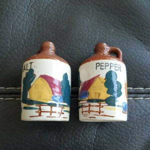May include: Salt and pepper shakers shaped like brown ceramic jugs with a white background. The shakers have a red and yellow house with a green tree on each side. The salt shaker says "SALT" and the pepper shaker says "PEPPER".