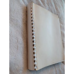 Pu&ograve; includere: Un quaderno a spirale con copertina color crema. Le pagine sono bianche e la rilegatura &egrave; argentata. Il quaderno &egrave; appoggiato su una superficie morbida e strutturata.