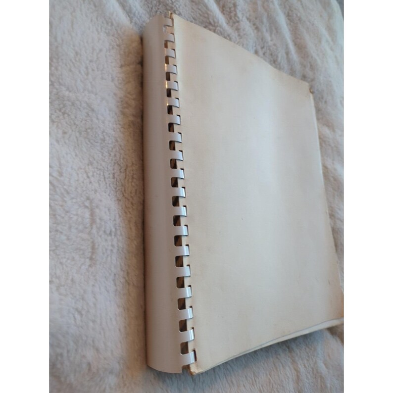 Pu&ograve; includere: Un quaderno a spirale con copertina color crema. Le pagine sono bianche e la rilegatura &egrave; argentata. Il quaderno &egrave; appoggiato su una superficie morbida e strutturata.