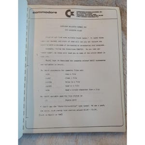 Pu&ograve; includere: Una pagina aperta di un manuale Commodore, con il logo Commodore in alto. Il testo include istruzioni sui file a cassetta, le istruzioni BASIC e lo stato dei file. La carta &egrave; ingiallita.