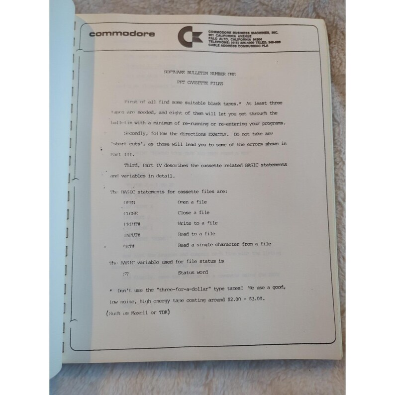Pu&ograve; includere: Una pagina aperta di un manuale Commodore, con il logo Commodore in alto. Il testo include istruzioni sui file a cassetta, le istruzioni BASIC e lo stato dei file. La carta &egrave; ingiallita.
