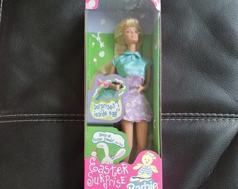 May include: A Barbie doll wearing a light blue and purple outfit with a pink Easter basket. The box features the text "Easter Surprise Barbie" and a warning about choking hazards.