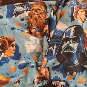 Può includere: Un tessuto blu e bianco a tema Star Wars con immagini di Chewbacca, Darth Vader, la Principessa Leia e R2-D2. Il tessuto ha uno sfondo blu con stelle bianche e le parole "Star Wars" stampate su di esso.